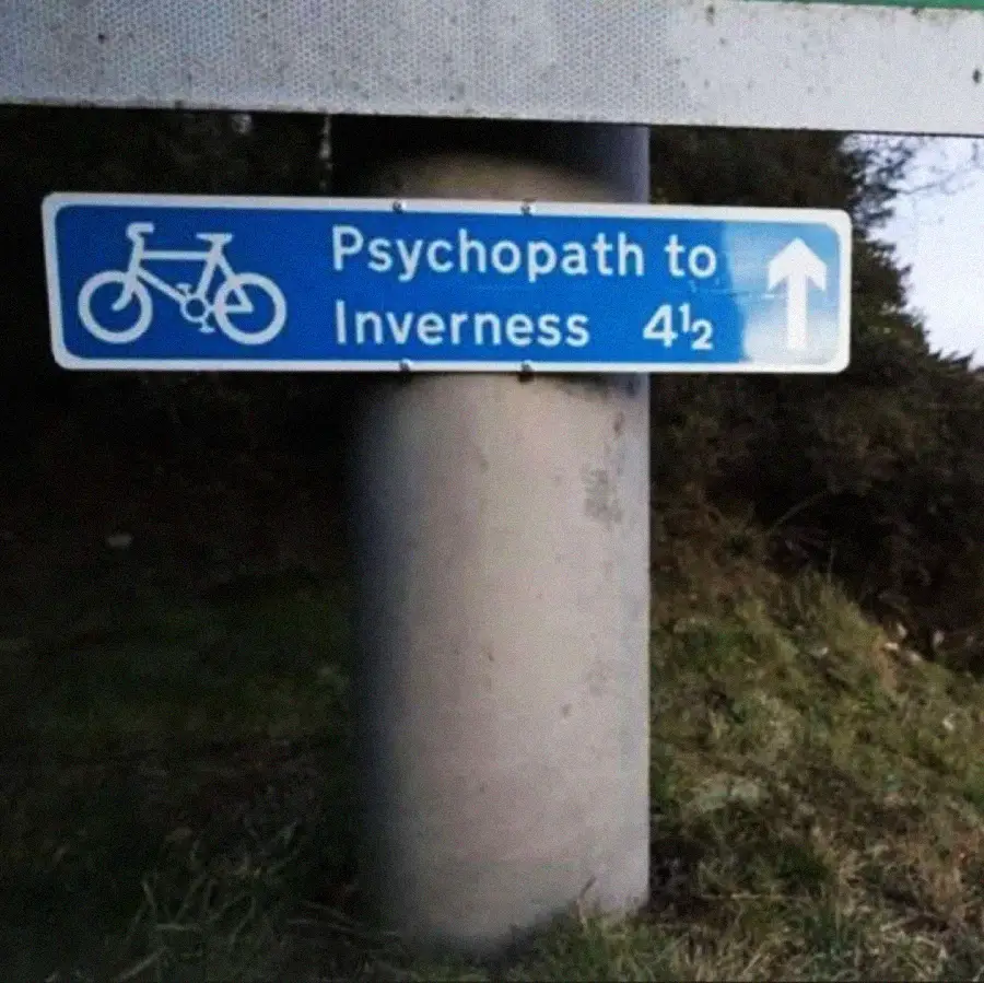 A blue road sign with a bicycle symbol reads, "Psychopath to Inverness 4½," with an arrow pointing right. The sign is attached to a concrete pole, and there is grass and shrubbery in the background.