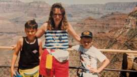 A woman and two young boys stand in front of a railing overlooking the Grand Canyon. The woman wears sunglasses, a striped tank top, and red shorts. The boys wear casual summer clothes and smile at the camera.