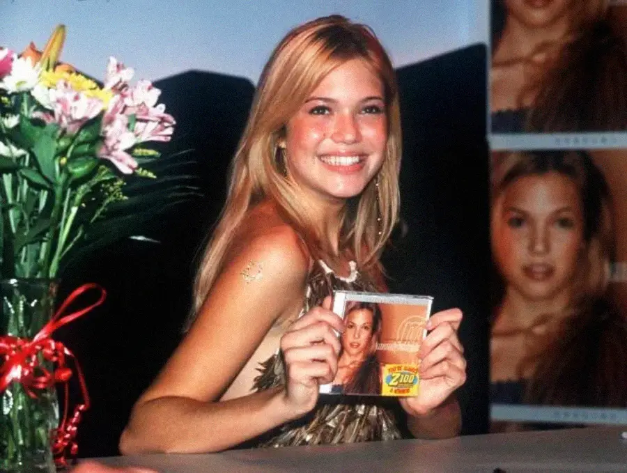 A young woman with long blonde hair smiles while holding a CD with her photo on the cover. She sits at a table with a bouquet of flowers, and a large poster of her face is visible in the background.