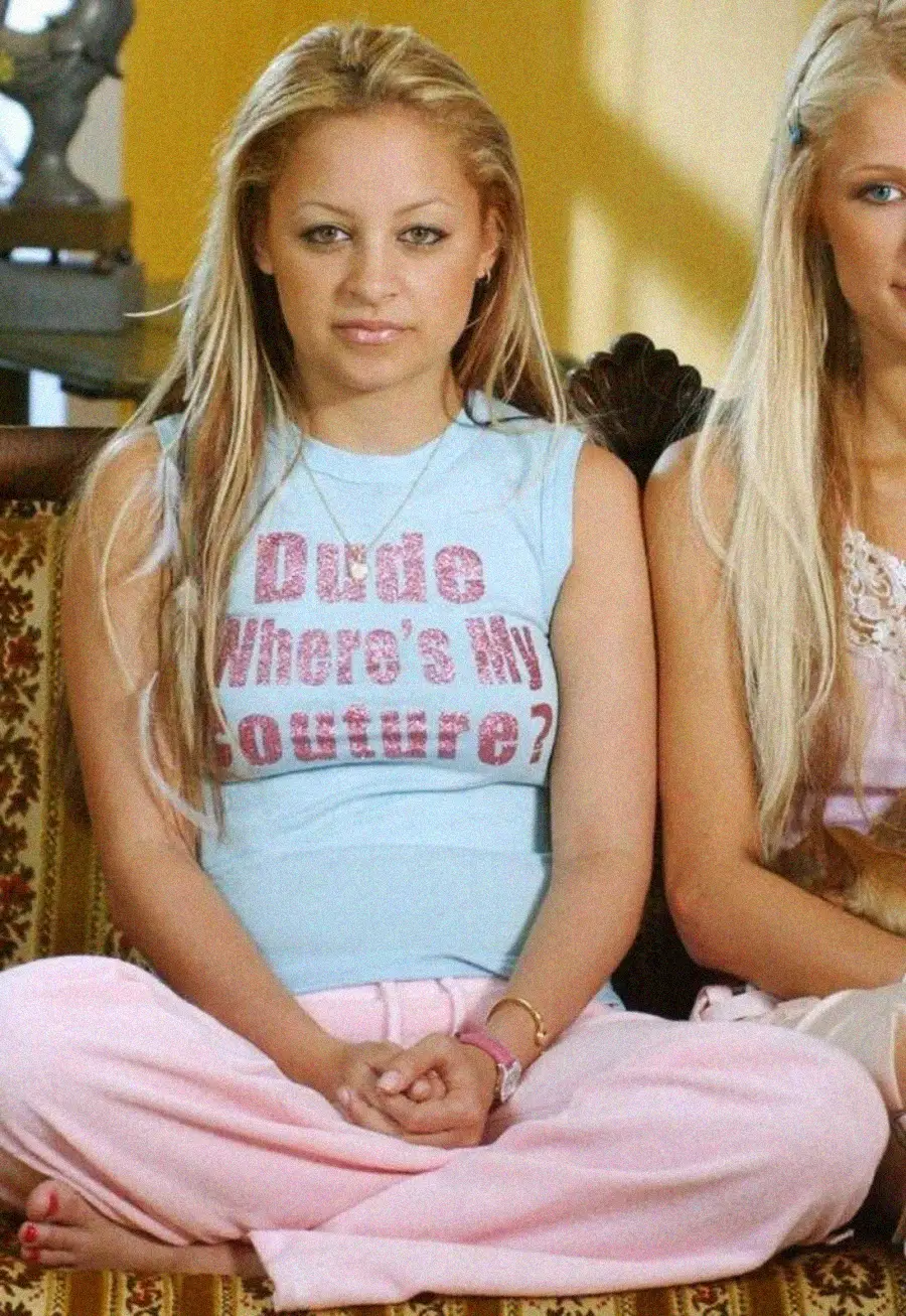 A woman with long blonde hair sits cross-legged on a patterned couch, wearing a light blue shirt that says "Dude Where’s My Couture?" in glittery pink letters and light pink pants. Another person sits beside her, partially visible.