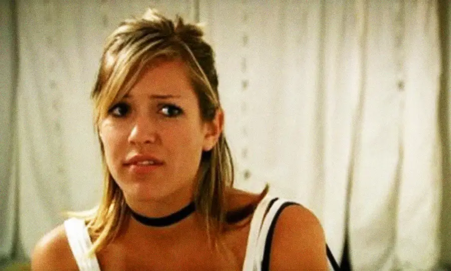 A young woman with blond hair, wearing a black choker and a white top, looks slightly uncomfortable or concerned while sitting indoors in front of white curtains.