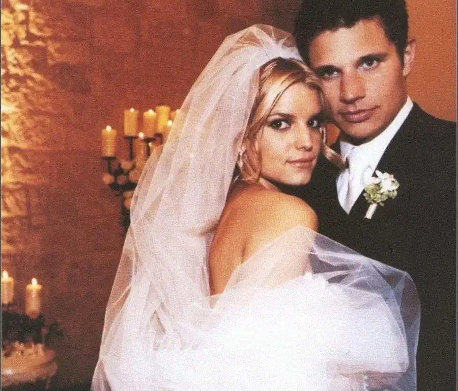 A bride in a white gown and veil embraces a groom in a dark suit and boutonnière. They pose closely together, looking at the camera, with candles softly glowing in the background.