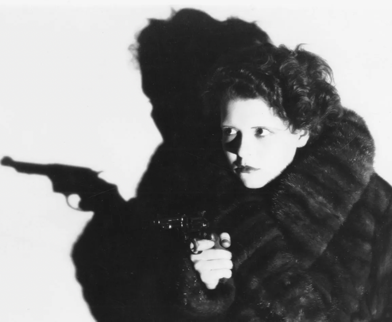 A woman in a fur coat holds a revolver, her face partially lit, with a dramatic shadow of herself and the gun cast on the white wall behind her.