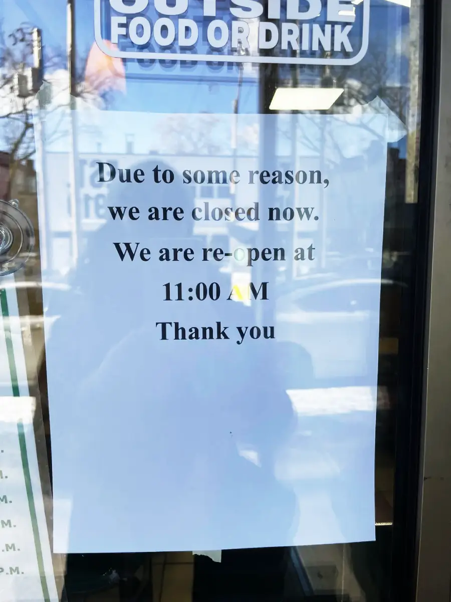 A printed sign on a glass door reads: "Due to some reason, we are closed now. We are re-open at 11:00 AM. Thank you," with reflections of buildings and trees visible on the glass.
