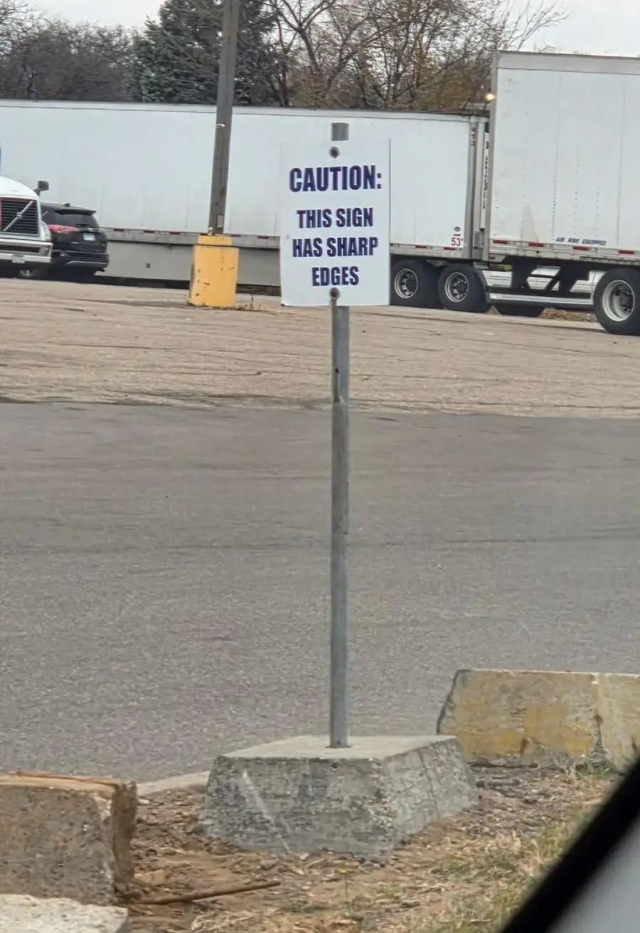 A metal sign in a parking lot reads "CAUTION: THIS SIGN HAS SHARP EDGES" in blue text. The sign is mounted on a metal pole set in a concrete block. Trucks and cars are parked in the background.