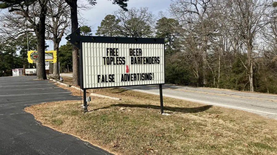A roadside marquee sign reads: "FREE TOPLESS BEER BARTENDERS FALSE ADVERTISING!!" with the words arranged so that "TOPLESS" and "BARTENDERS" are on separate lines, creating a humorous message.