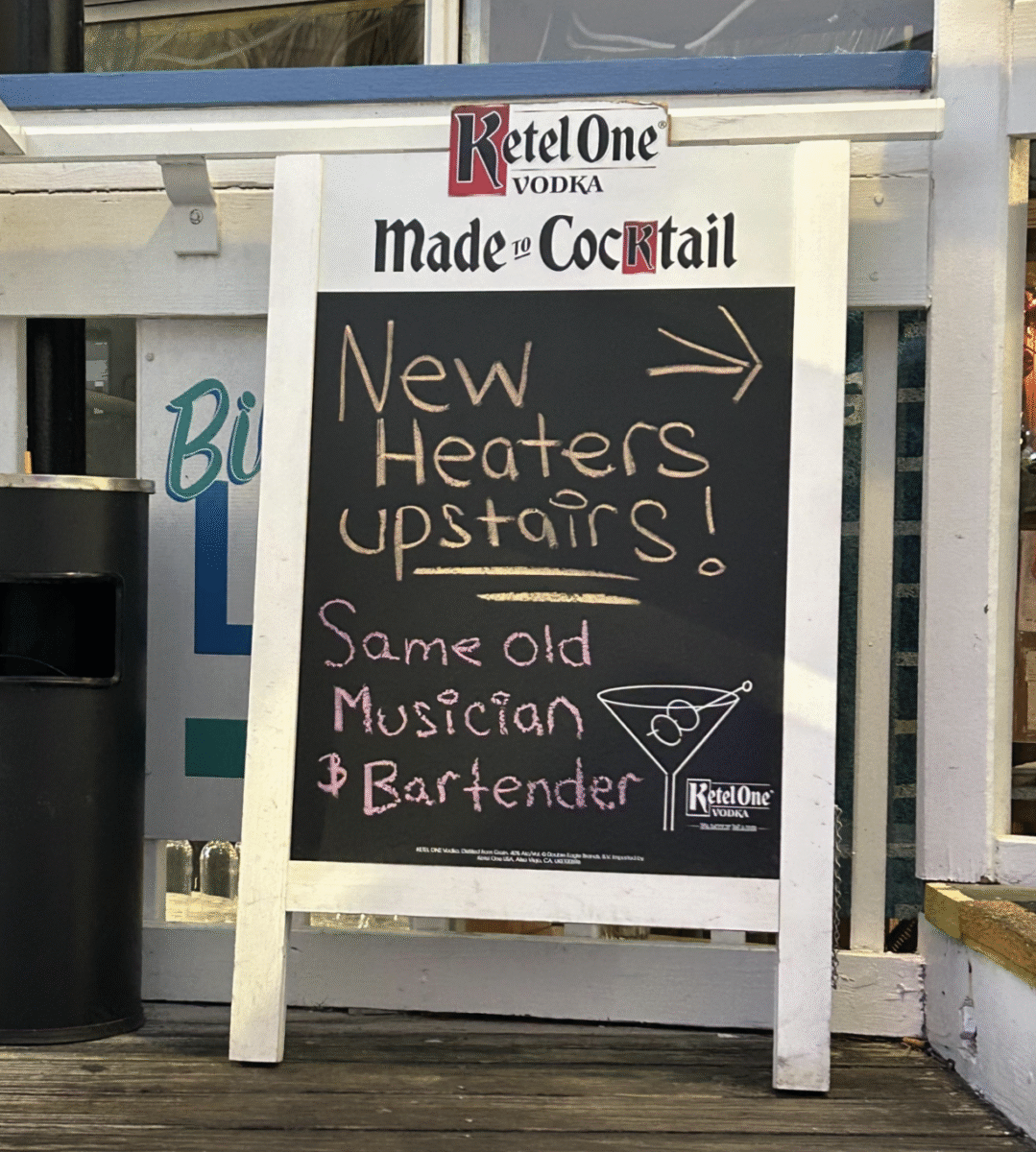 A chalkboard sign outside a bar reads: "New heaters upstairs! Same old musician & bartender." The sign features a Ketel One Vodka logo and a simple martini glass drawing.