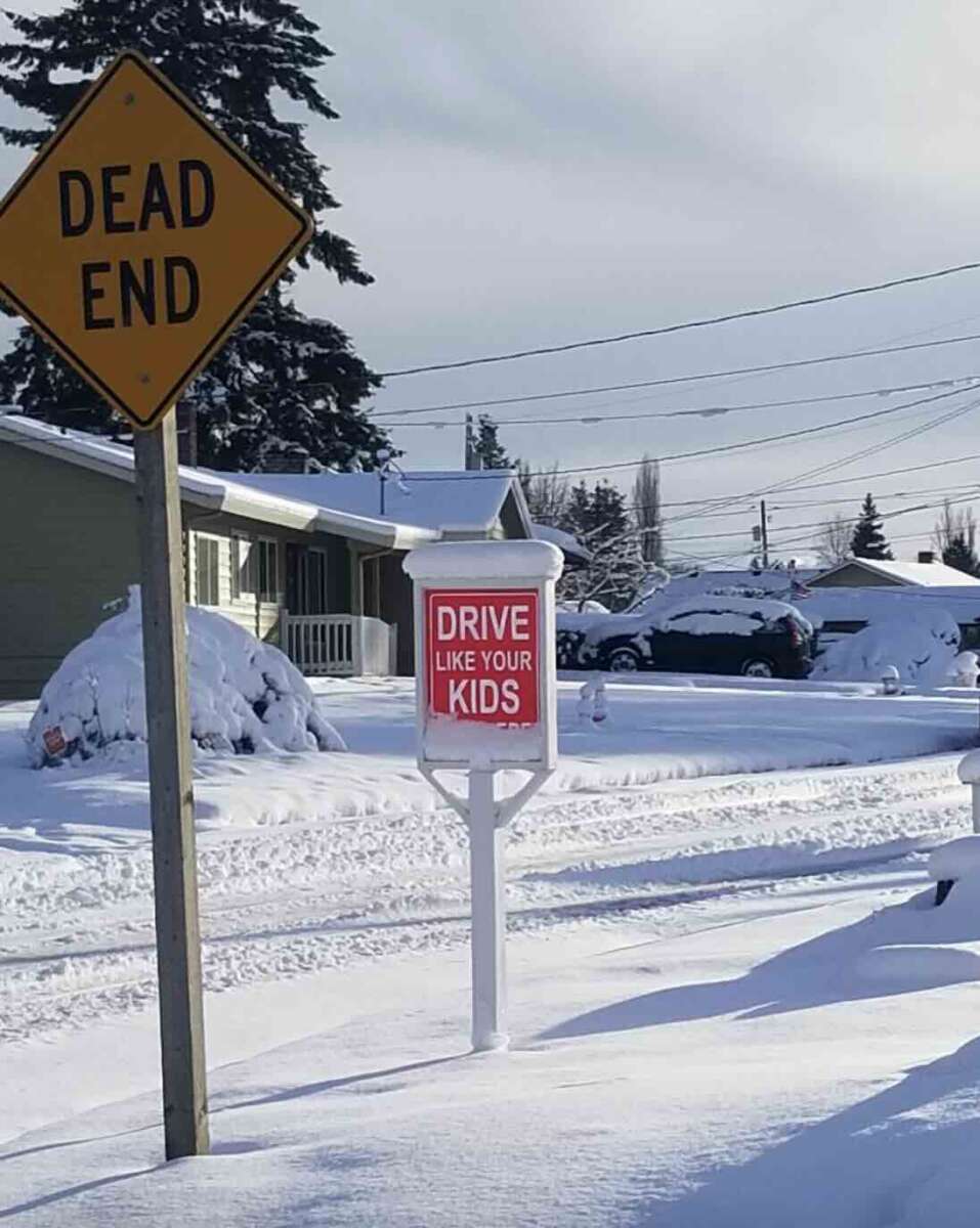 A "DEAD END" street sign and a sign reading "DRIVE LIKE YOUR KIDS LIVE HERE" stand beside a snow-covered residential street lined with houses and parked cars. Thick snow blankets the ground and trees.
