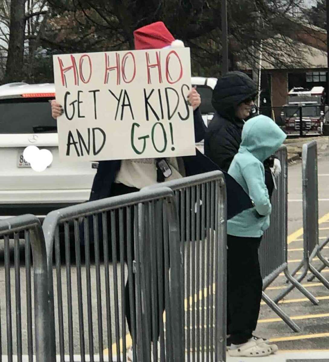 A person in a Santa hat holds a sign reading “HO HO HO GET YA KIDS AND GO!” while standing beside two people, all near metal barricades in a parking lot. Trees and cars are visible in the background.
