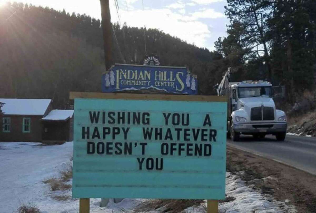 A roadside sign at Indian Hills Community Center reads, "WISHING YOU A HAPPY WHATEVER DOESN'T OFFEND YOU," with a truck driving on the road beside it and snowy ground in the background.