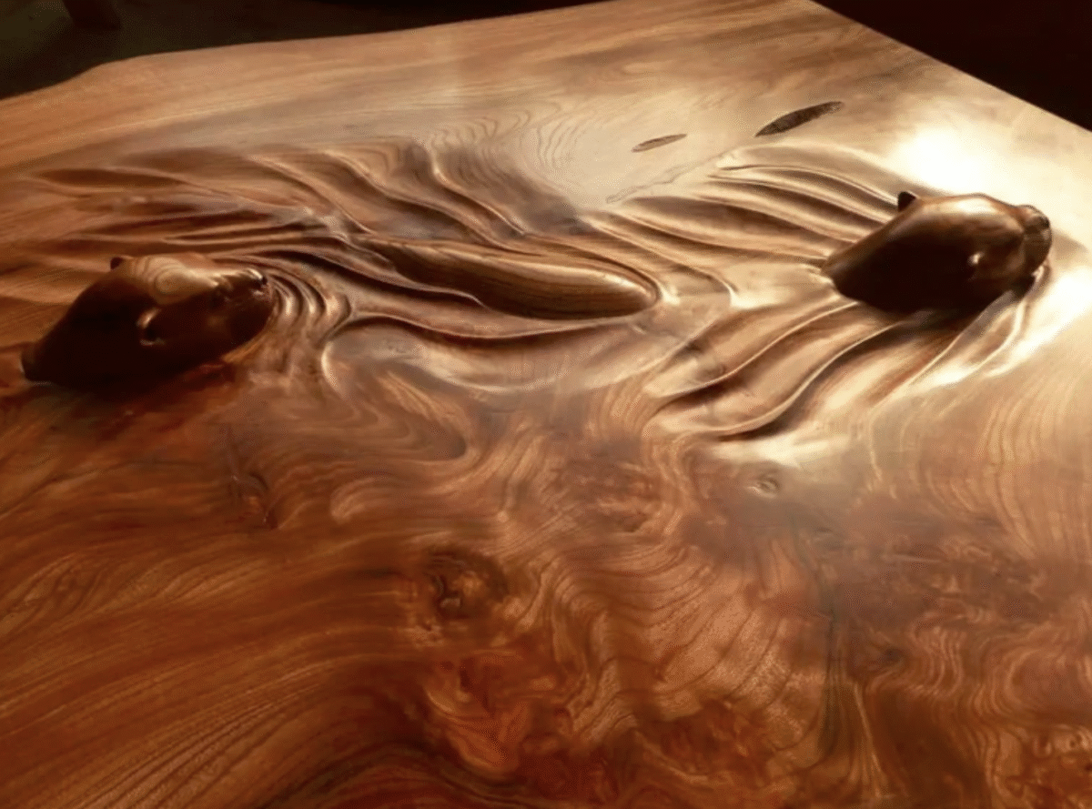 A polished wooden surface sculpted to resemble two otters swimming, with their heads and bodies emerging smoothly from the flowing wood grain.
