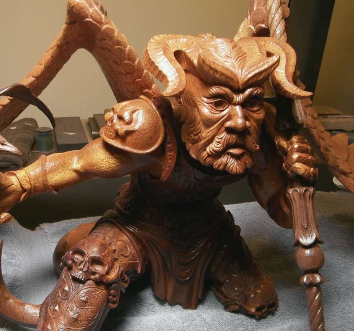 A detailed wooden sculpture of a demon-like figure with horns, wings, and an intense expression, holding a staff. Skulls are carved into its armor and knee, showcasing intricate craftsmanship.