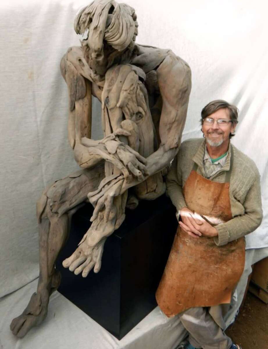 A smiling artist wearing glasses and a brown apron sits beside a large, expressive, abstract clay sculpture of a seated human figure with elongated limbs. Both are positioned against a neutral backdrop.