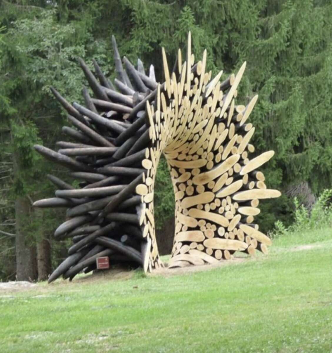 A large outdoor sculpture made of stacked logs forms an archway, with one side featuring light-colored smooth wood and the other side built from dark, jagged, charred logs, set against a backdrop of green trees.