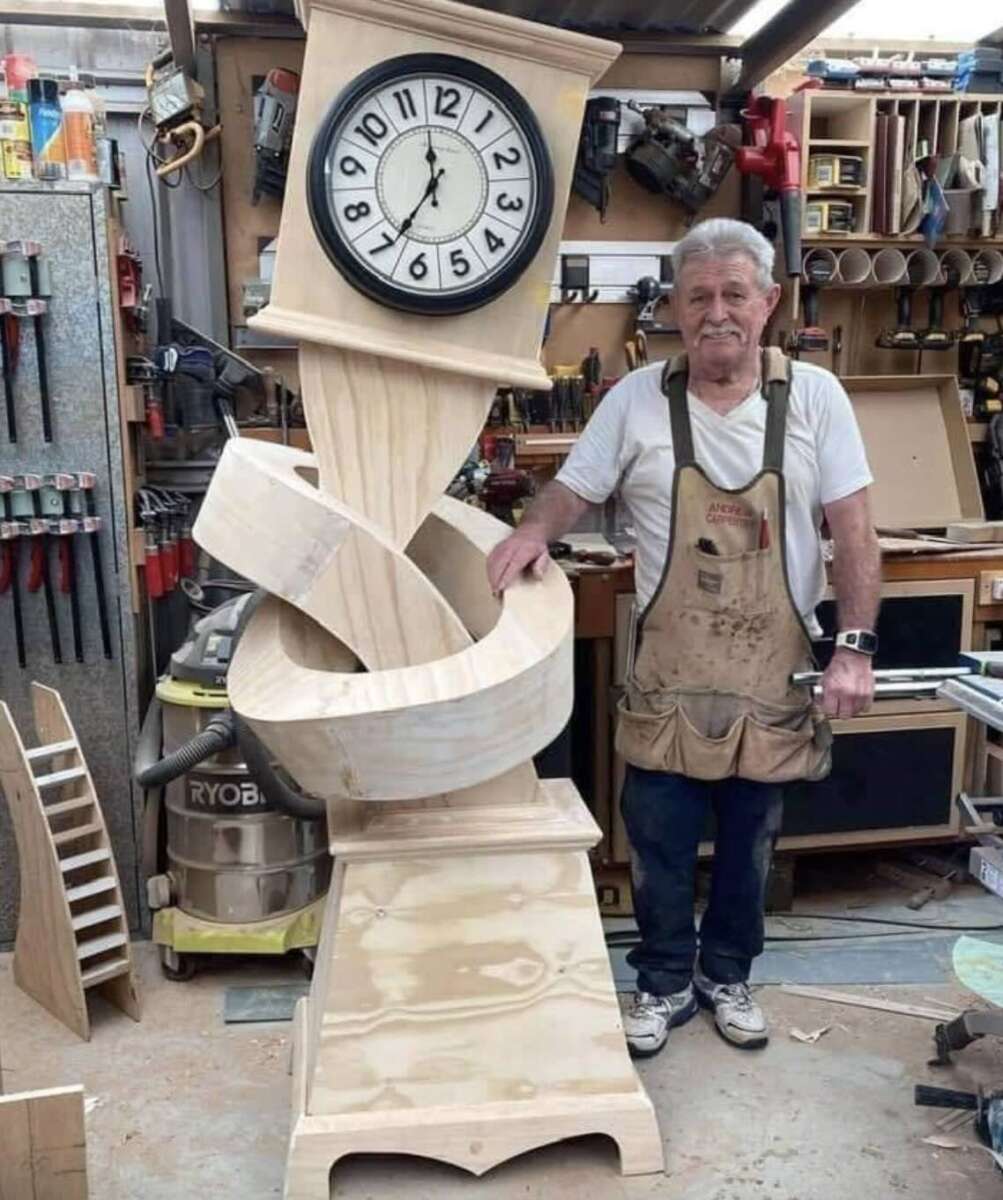 An older man in a workshop stands beside a large, unfinished wooden grandfather clock with a twisted, spiral design. Tools and woodworking equipment are visible in the background.