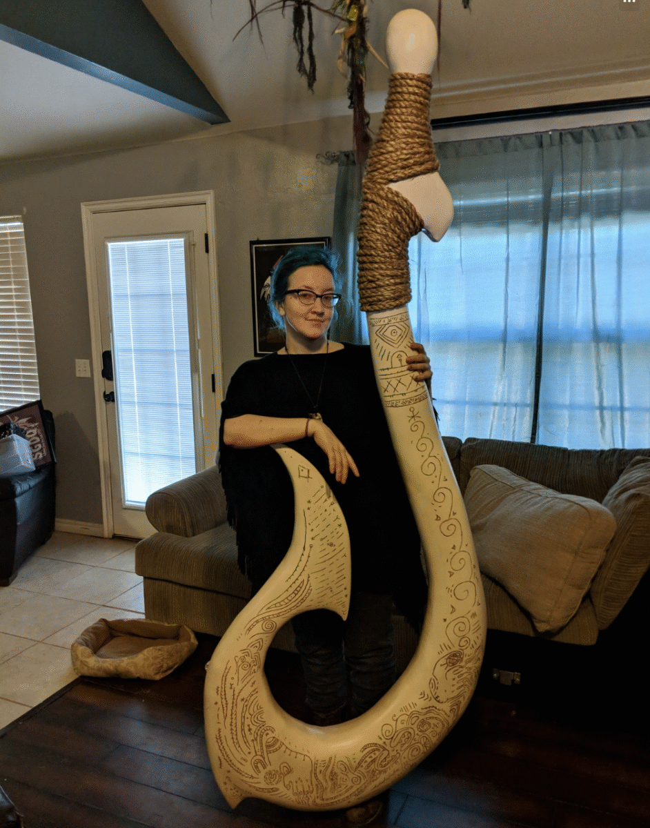 A person with blue hair stands indoors, holding a large, ornate, cream-colored hook prop decorated with intricate patterns and wrapped with brown twine at the handle. They are smiling and standing beside a couch.