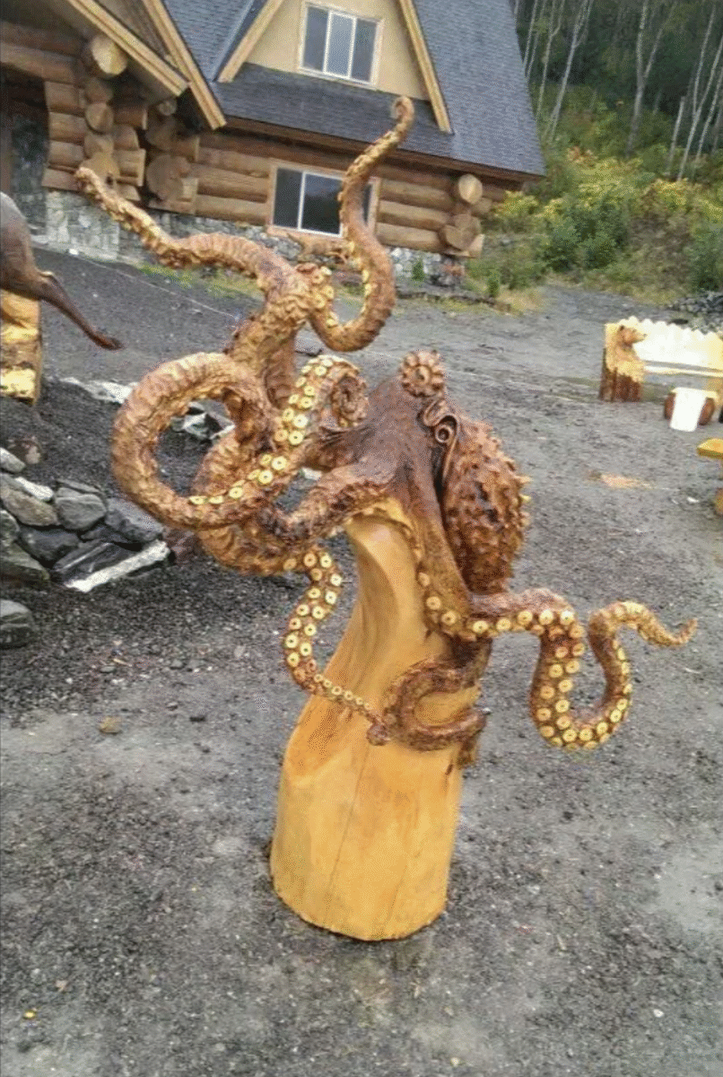 A detailed wooden sculpture of an octopus with extended tentacles stands outdoors in front of a log cabin surrounded by trees.