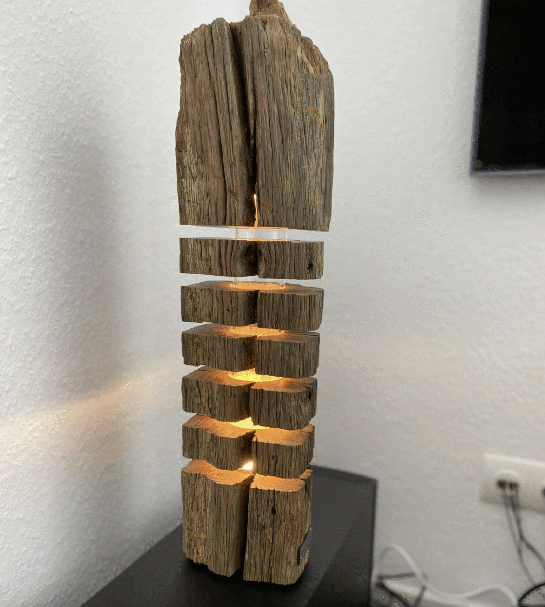 A rustic, vertical wooden lamp with horizontal slits emitting warm yellow light stands on a black surface against a white wall. The lamp is made from weathered wood, giving it a natural, textured appearance.