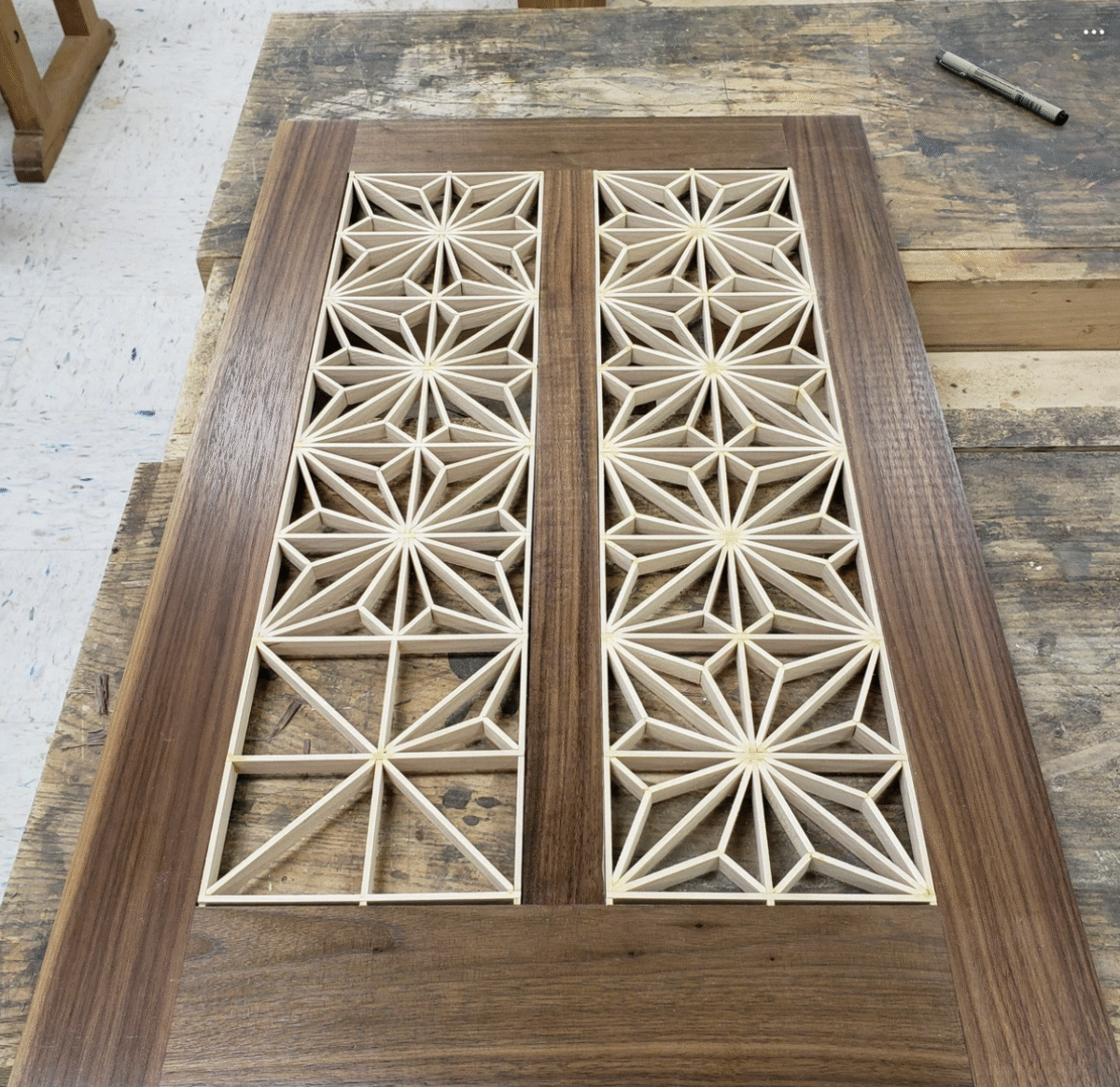 A wooden panel with two rectangular sections filled with intricate geometric patterns made from thin, light-colored wood, set on a workbench in a woodworking shop.