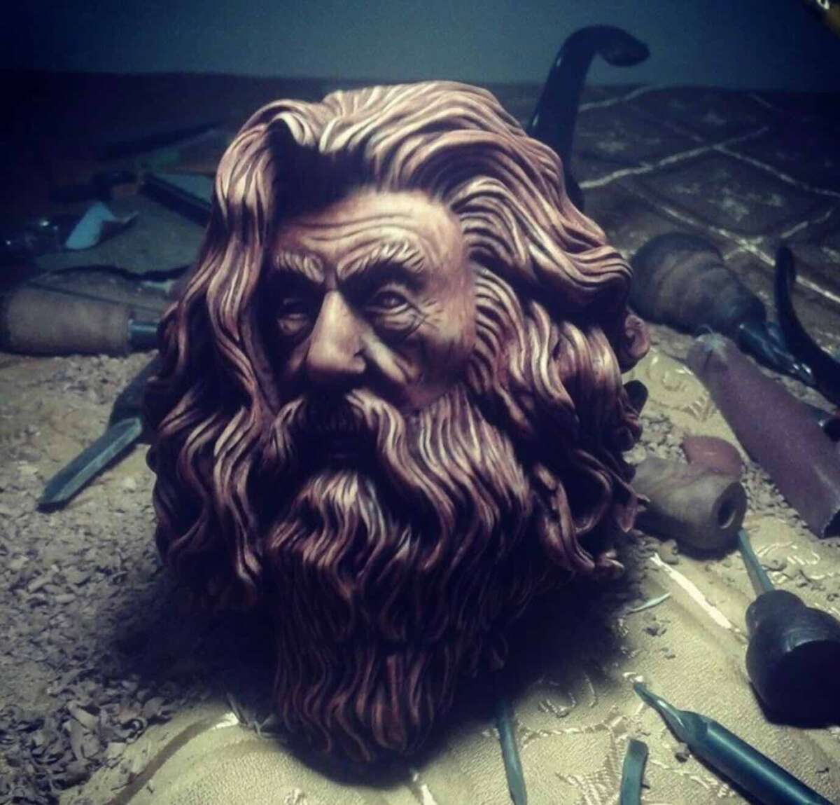 A detailed wooden sculpture of a bearded man's head with flowing hair is surrounded by carving tools and wood shavings on a workbench.