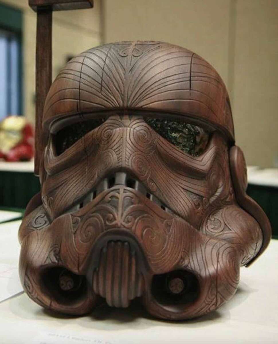 A Stormtrooper helmet designed to look like carved wood, featuring intricate, swirling patterns and engraved details, displayed on a tabletop at an indoor event.