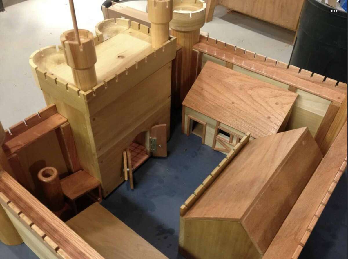 A detailed wooden model of a medieval castle, featuring towers, a gatehouse with an open door, walls with battlements, and inner buildings arranged on a blue-gray surface.