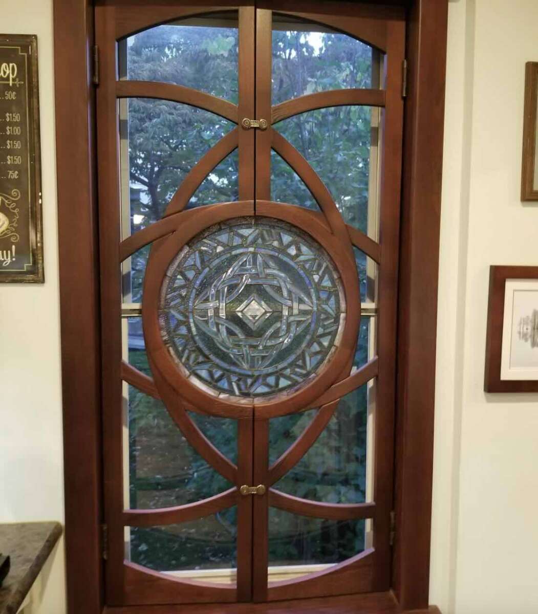 A wooden-framed window with an intricate circular stained glass design in the center, featuring geometric patterns. Outside, green trees are visible through the glass. Framed art hangs on the wall beside the window.