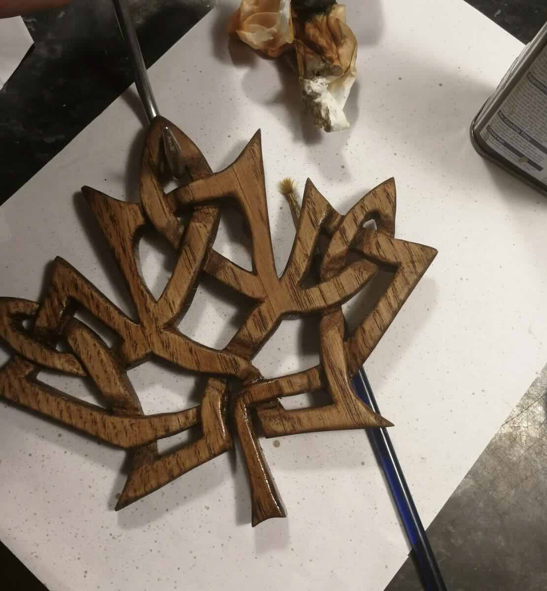 A hand applies varnish to a wooden, Celtic knot-style maple leaf decoration resting on white paper, with a paintbrush, paper towel, and a can off to the side.