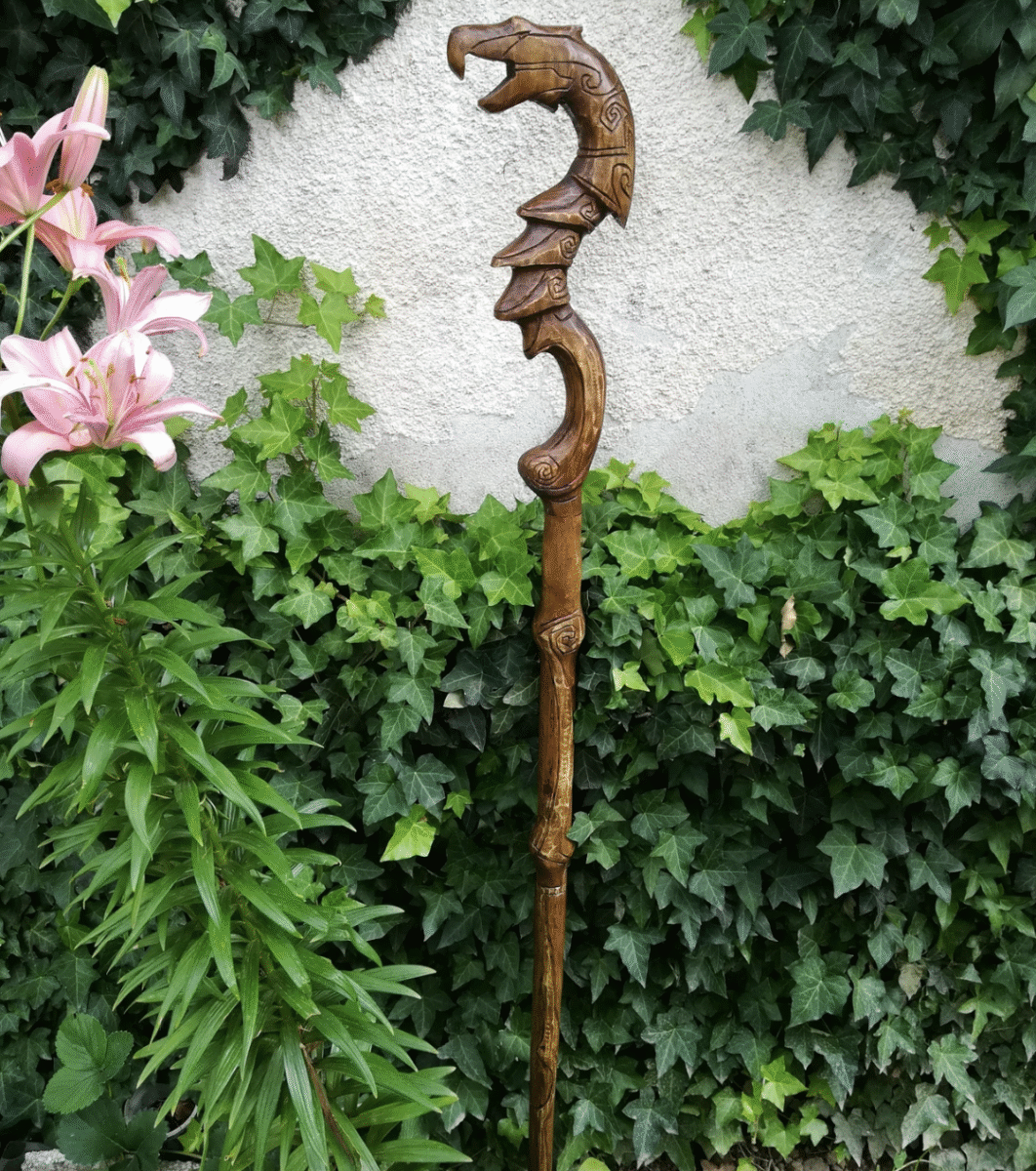 A carved wooden staff with a dragon head design at the top stands upright among lush green ivy and pink lilies, in front of a textured beige wall.