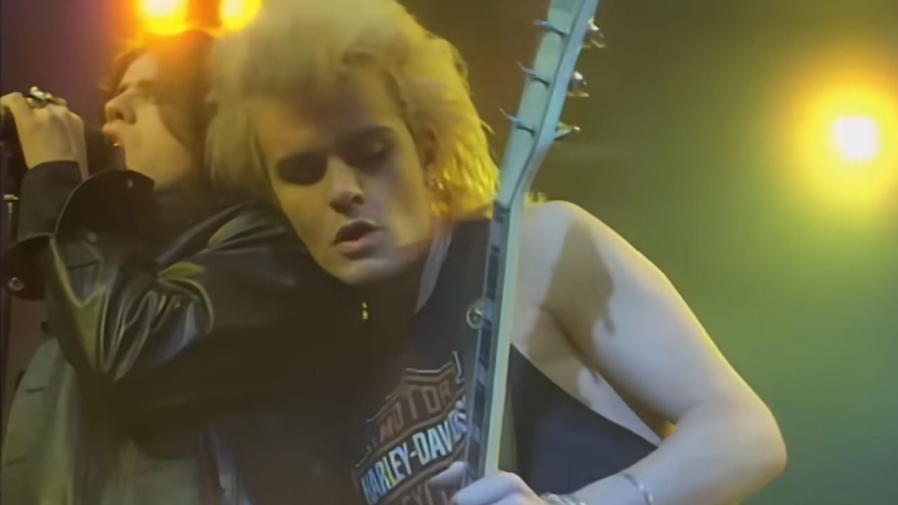 Two musicians perform on stage under bright yellow lights. One sings into a microphone while the other plays guitar, wearing a sleeveless Harley-Davidson shirt and leaning in closely.