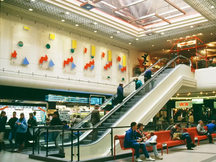 People ride an escalator inside a brightly lit shopping mall with colorful abstract art on the wall, stores in the background, and shoppers sitting on red benches in the foreground.