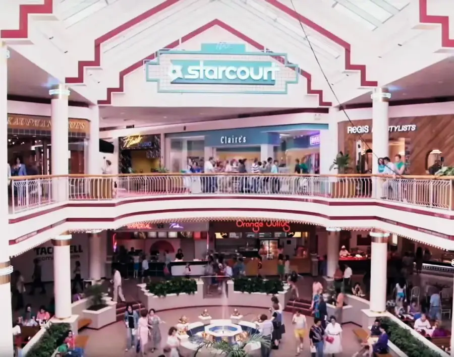 A busy shopping mall with two levels, featuring a large "Starcourt" sign above stores like Claire’s and Orange Julius, with people walking, shopping, and gathering around a central fountain.