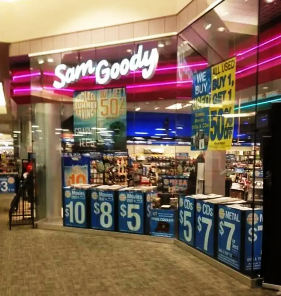 A Sam Goody store with bright neon signage, blue sale posters in the windows advertising deals on movies and CDs, and shelves of music and movies visible inside.