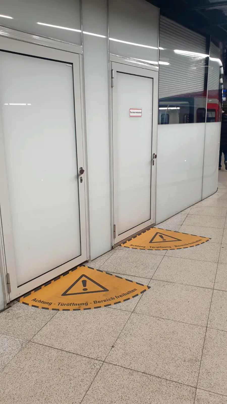 Two white doors at a train station with yellow warning signs on the floor in front of them, alerting about door openings. A train is visible on the right side of the image.