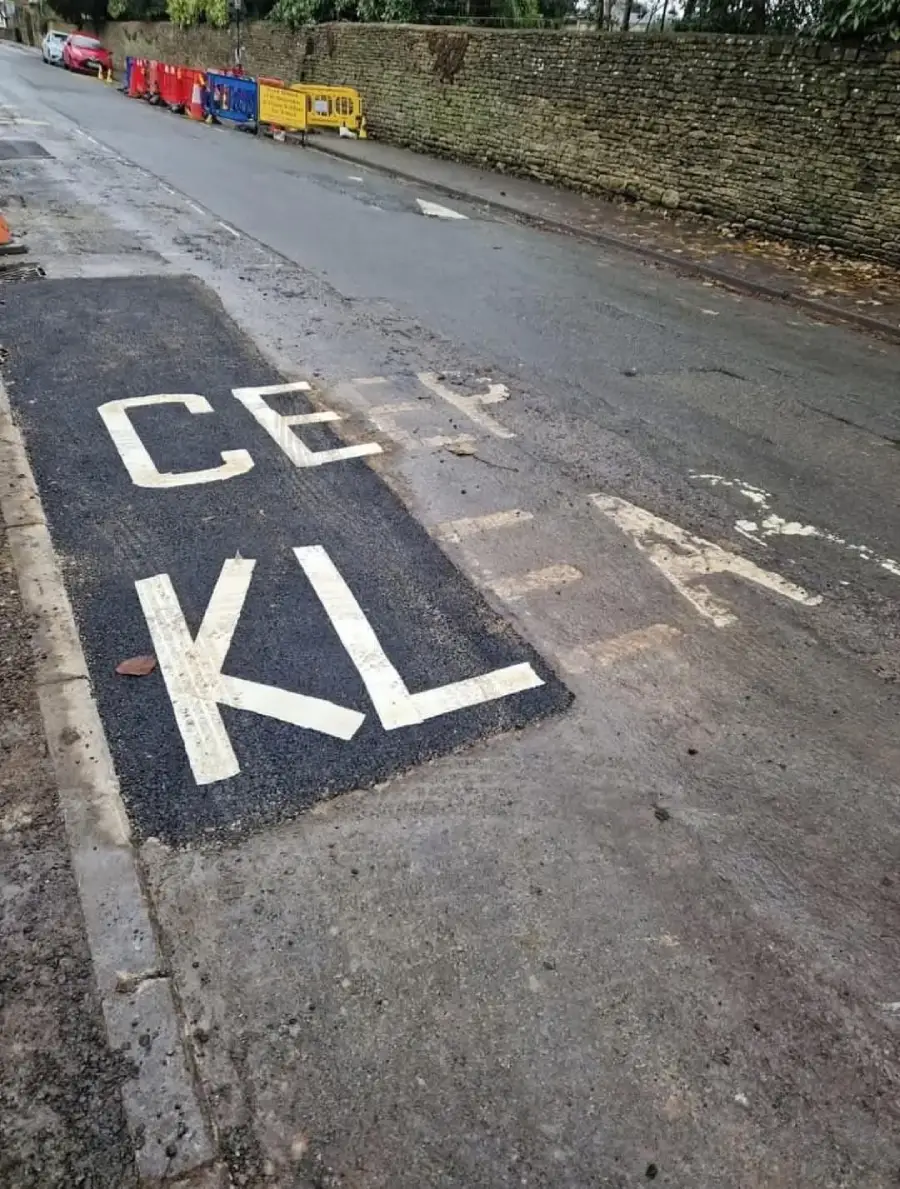 Road markings saying "KEEP CLEAR" are partially repainted as "CEP KLEA" on fresh asphalt while the faded original text is still visible beneath. Barriers line the road, and a stone wall runs alongside.