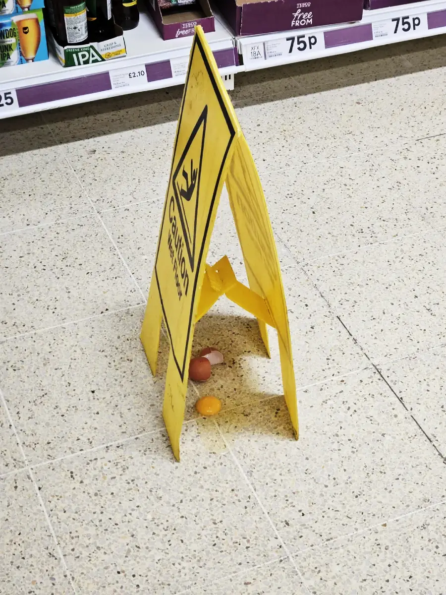 A yellow caution wet floor sign stands over a broken egg on a supermarket floor, with some spilt egg visible beneath the sign. Shelves with various products and price labels are in the background.