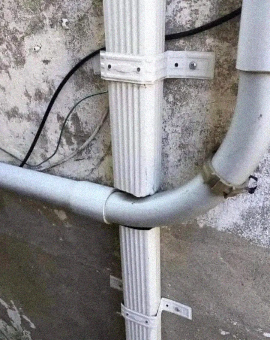 A white drainpipe is blocked by a horizontal gray pipe that passes directly through the downspout, preventing water from flowing downward. The pipes are mounted on a weathered, textured concrete wall.
