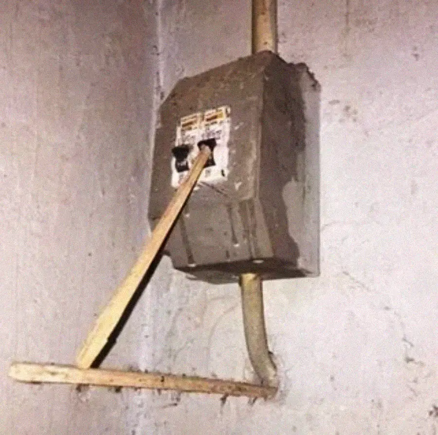 A wooden stick is propping up the switches of an old, dirty electrical box mounted on a concrete wall, creating a makeshift way to keep the switches in the "on" position.