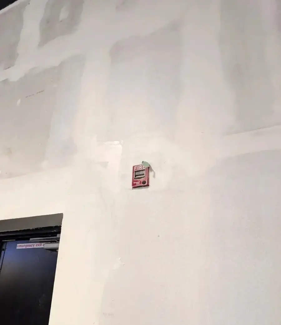 A fire alarm pull station is mounted unusually high up on a white wall, far out of normal reach. Part of a black door frame is visible at the bottom left of the image.