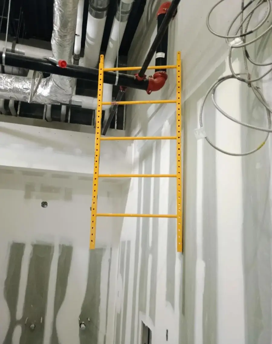 A yellow ladder is mounted vertically on a wall in an unfinished room, leading up to exposed pipes and ducts on the ceiling. The walls are partially covered with drywall and wiring is visible.