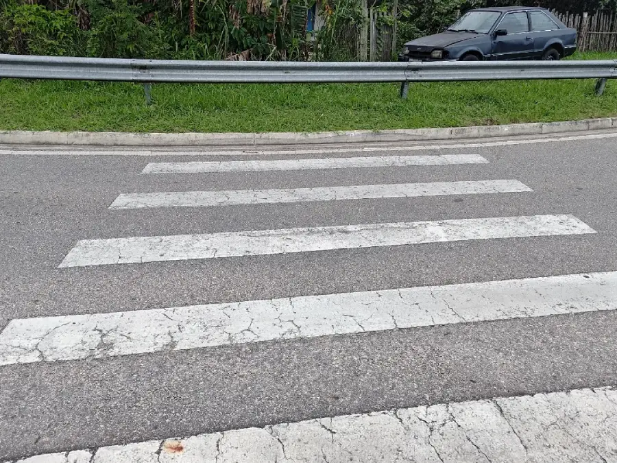 A crosswalk with faded white stripes on a gray road leads to a grassy area with a metal guardrail and an old parked car in the background.