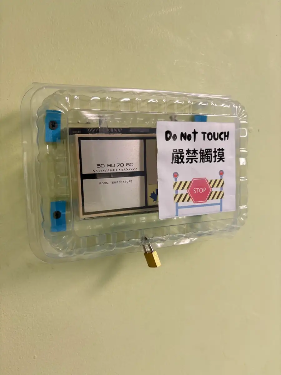 A thermostat covered by a clear plastic container, secured with a small padlock. A sign on the cover reads "DO NOT TOUCH" in English and Chinese, with a stop symbol. The setup is mounted on a light yellow wall.