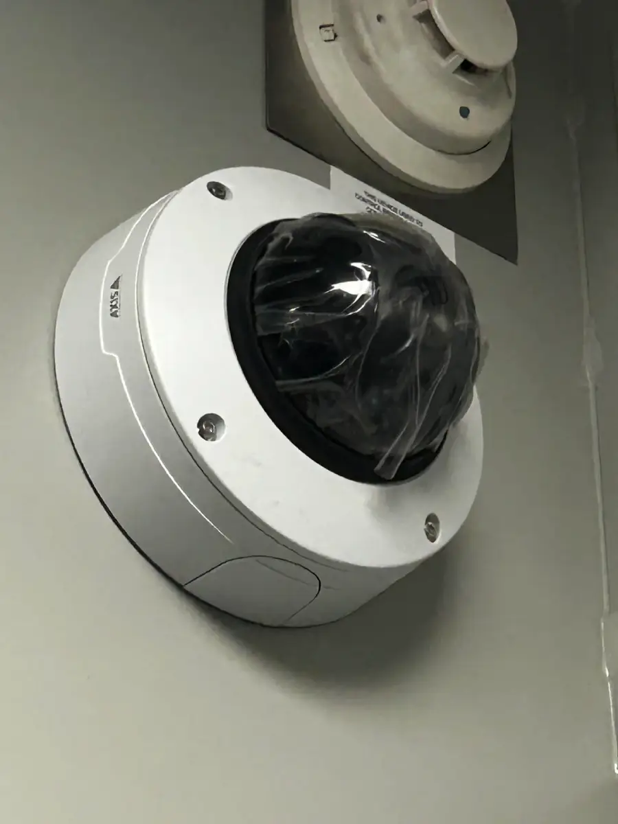 A security camera mounted on a wall with its black dome still covered in protective plastic, next to a smoke detector above it.