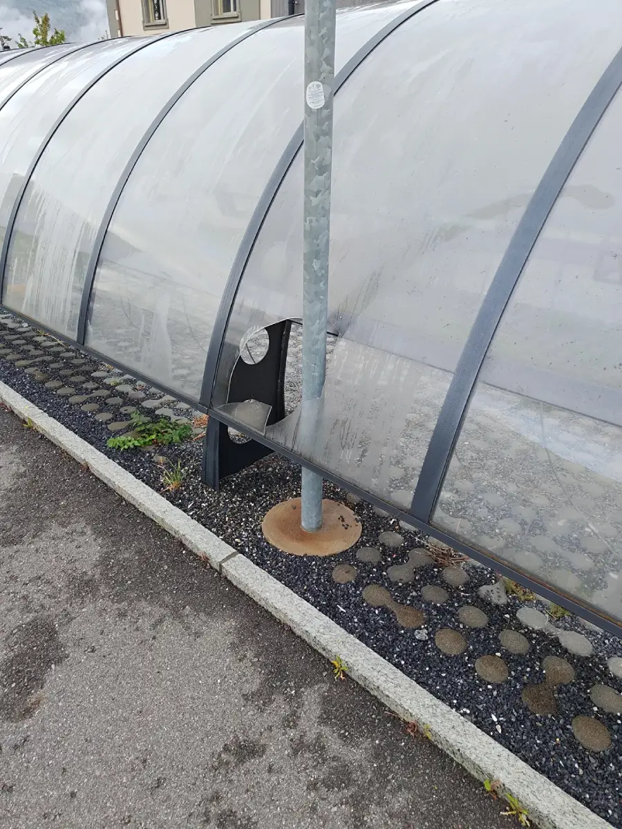 A metal pole goes through the side of a transparent plastic shelter, breaking the panel, with concrete and paving stones on the ground around it.