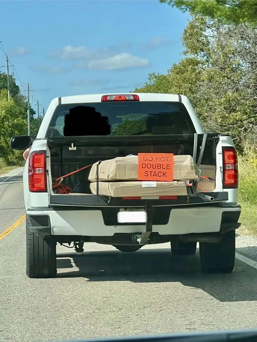 A white pickup truck drives with its tailgate down, carrying a large, wrapped package secured with straps. A bright orange sign on the package reads "DO NOT DOUBLE STACK.