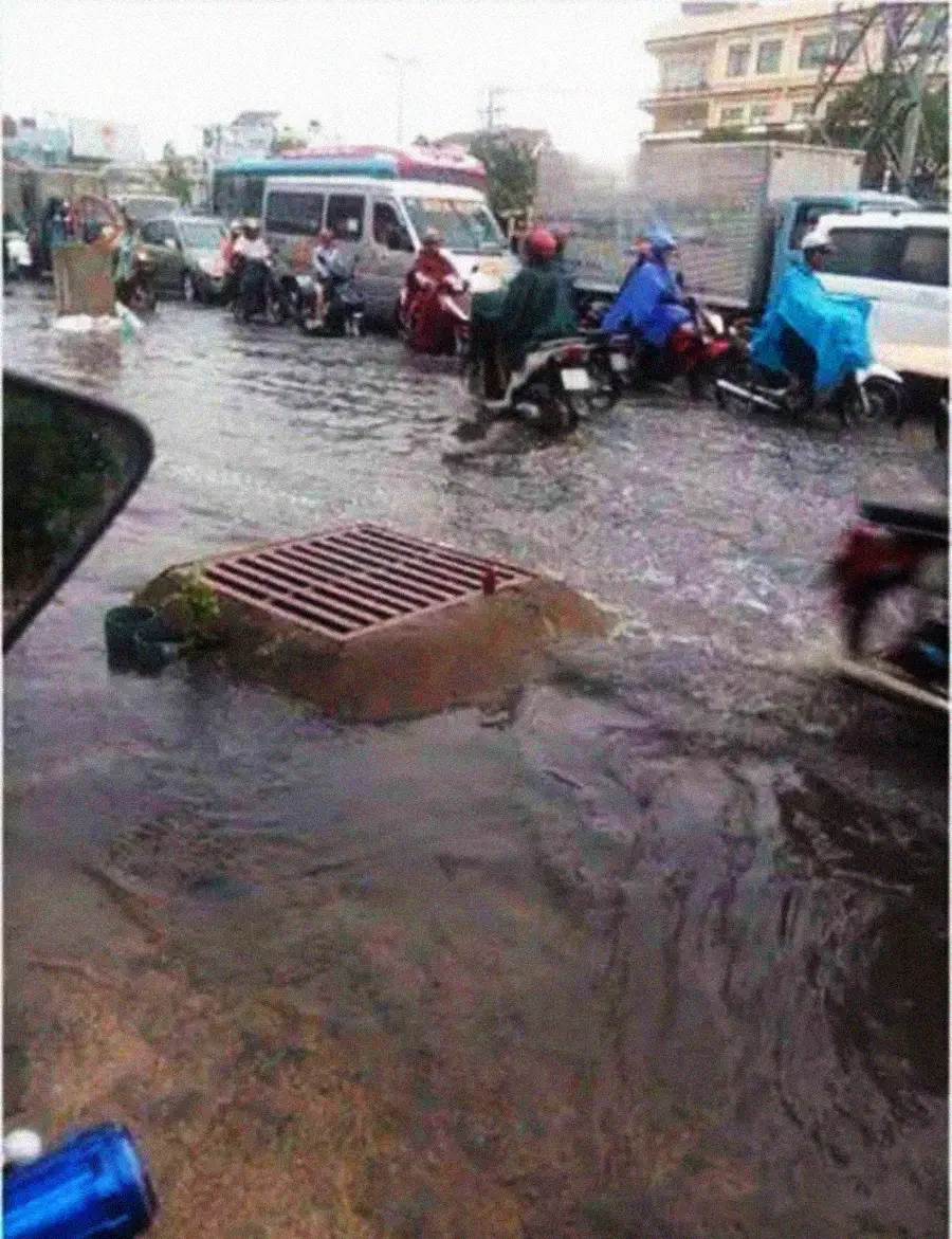 Flooded street with cars and motorcyclists driving through deep water; a submerged drain cover is visible in the center, creating a potential hazard.