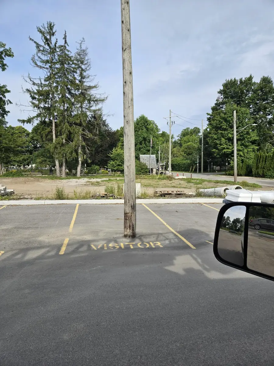 A wooden utility pole stands in the middle of a "VISITOR" parking space in an otherwise empty parking lot, with trees and construction materials visible in the background.