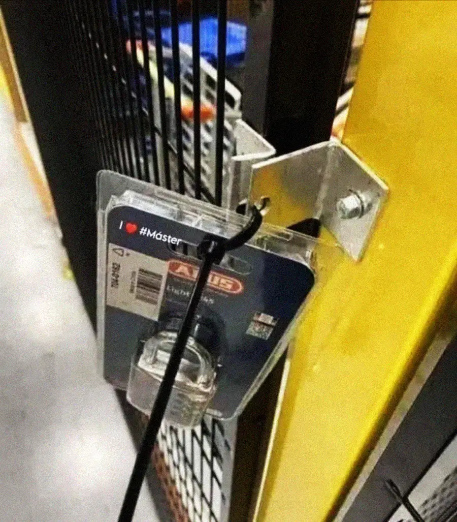 A packaged padlock is secured to a fence with a cable, still in its plastic packaging, instead of being used to lock the gate itself.