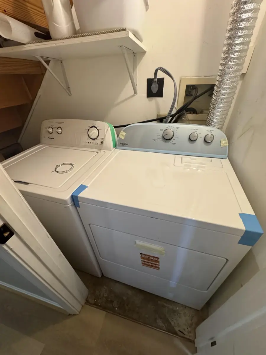 A small laundry room with a white washing machine and dryer side by side, both with blue tape on the edges. Shelves with containers are above, and a vent hose is attached to the dryer.