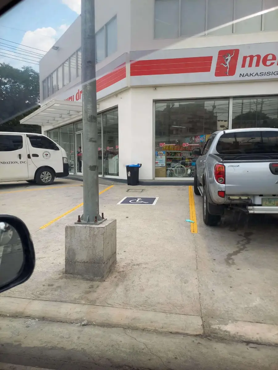 A concrete post blocks a parking space marked with a wheelchair-accessible symbol in front of a store, making the designated accessible spot unusable. Two vehicles are parked nearby.